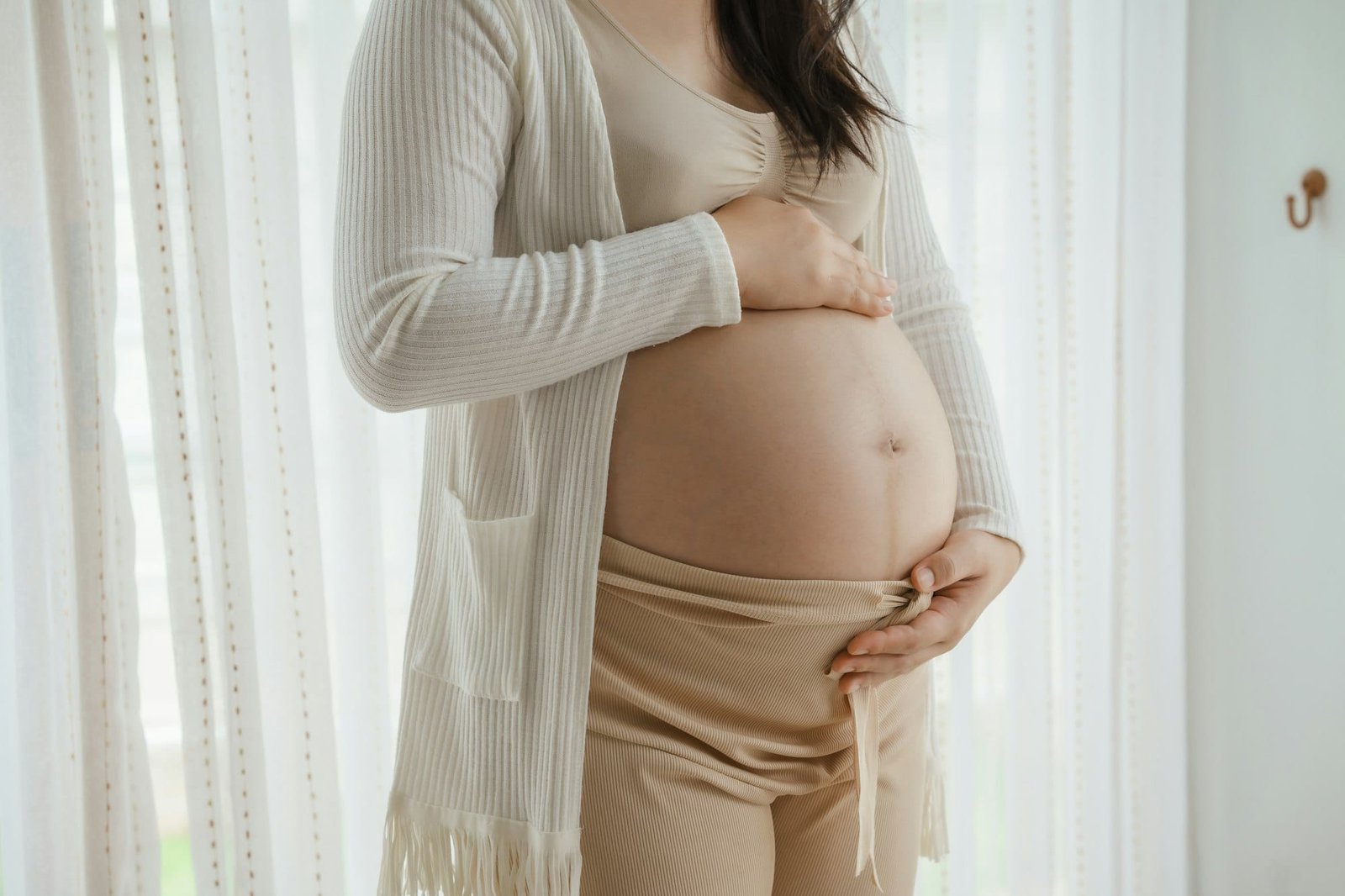 An Asian woman stands while pregnant with her first child, placing her hand on her stomach. She is